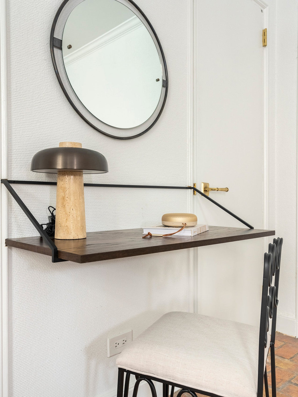 Dark wood floating desk with a travertine table lamp and a round wall mirror in a minimalist interior setting.