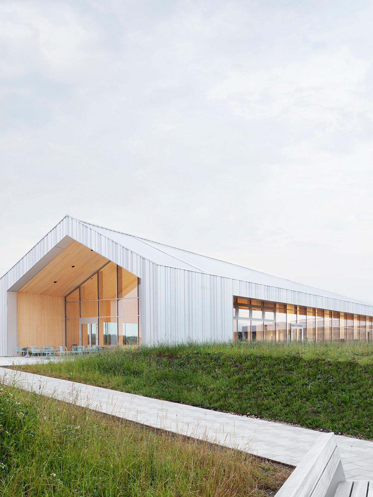 Minimal white metal-clad pavilion with pitched roof, large glass facade and warm wooden interior, outdoor chairs on concrete