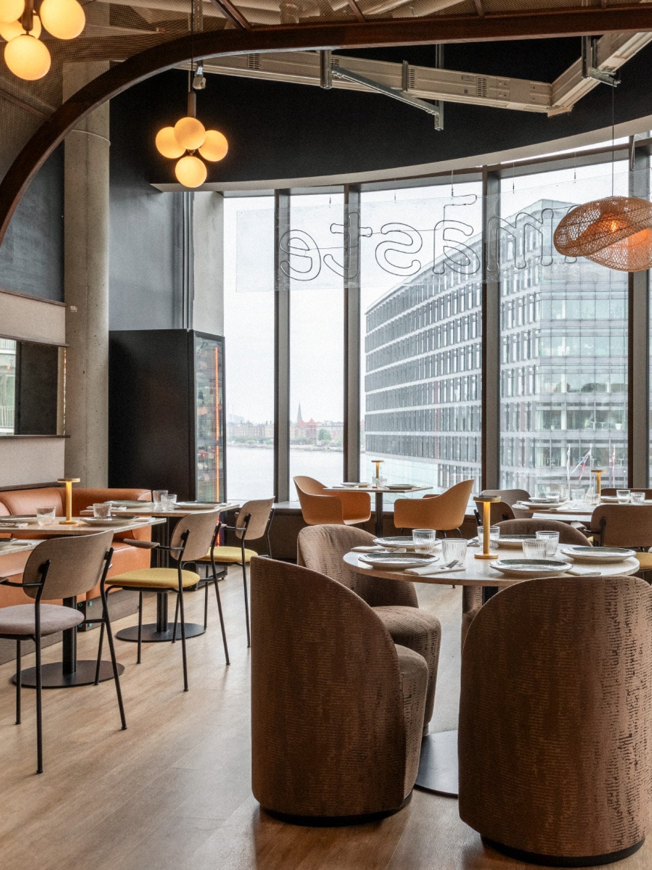 Modern restaurant interior with brown upholstered chairs, wooden tables set with glassware, globe pendant lights and city