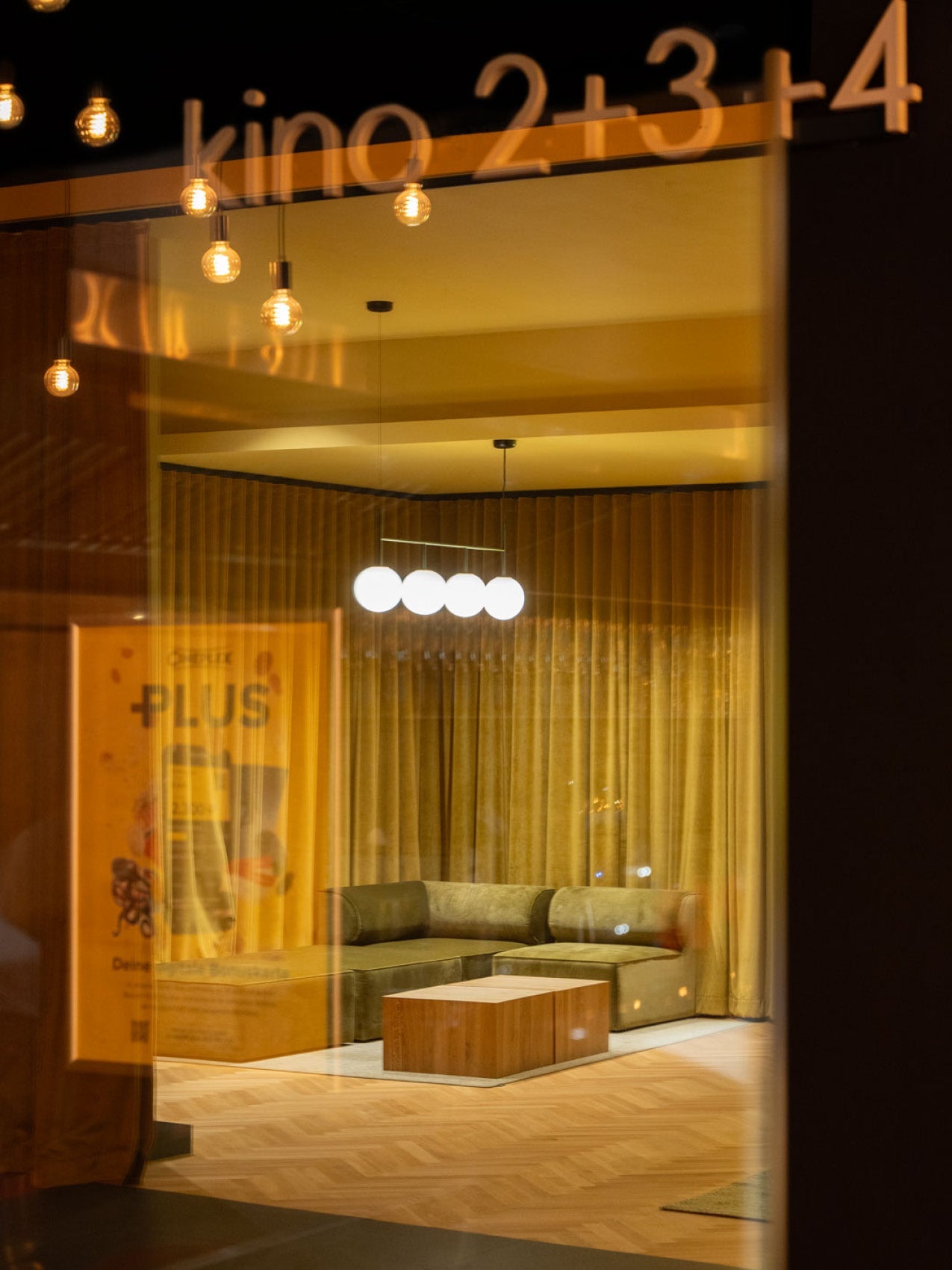 Interior showroom view through glass showing olive-green velvet L-shaped sofa, wooden cube coffee table and pendant globe
