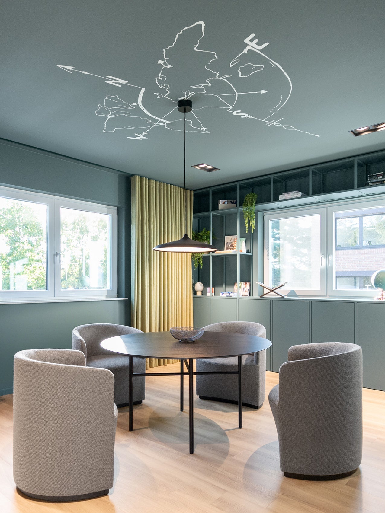 Dining nook with round dark wood table, four grey upholstered barrel chairs, pendant light and teal built-in shelving.