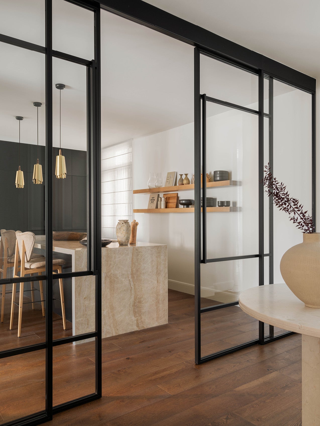 Interior view of black-framed glass sliding doors opening to a neutral dining area with marble island, wooden shelves and