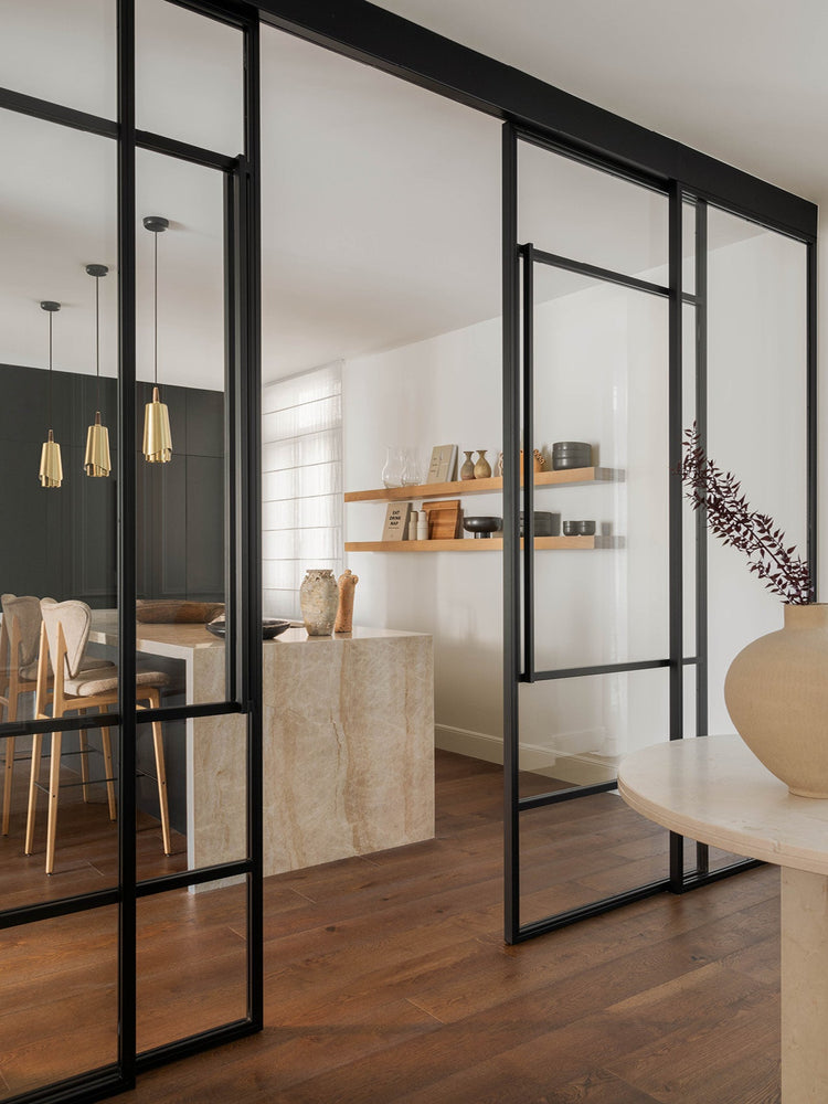 Interior view of black-framed glass sliding doors opening to a neutral dining area with marble island, wooden shelves and