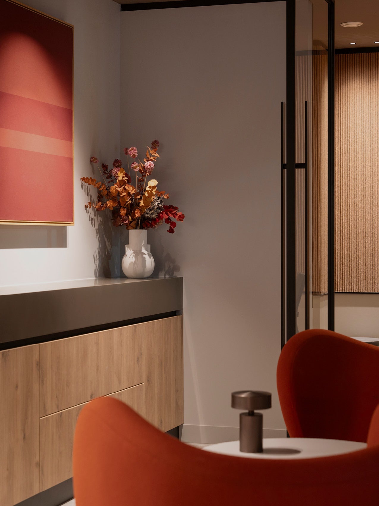 Minimal interior with a wooden sideboard, white ceramic vase of dried orange and red foliage, and a curved terracotta