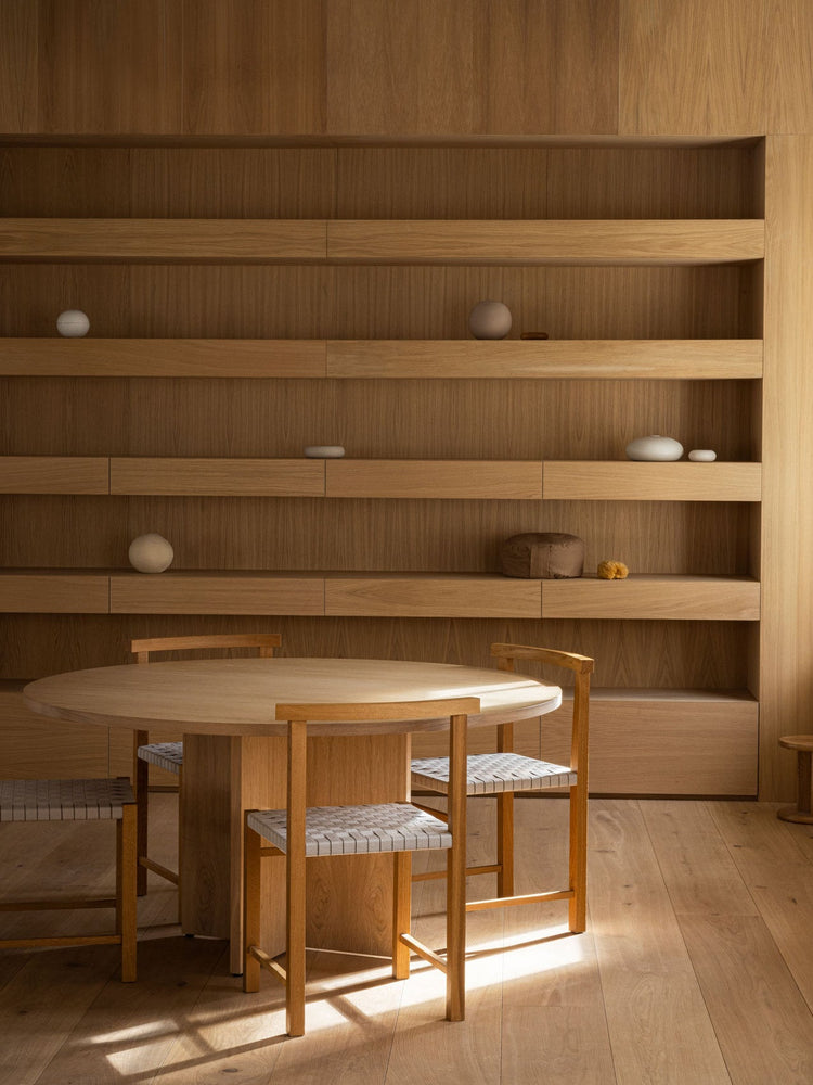Minimal oak dining table and woven-seat chairs in front of built-in oak shelving with neutral ceramic and fabric decor.