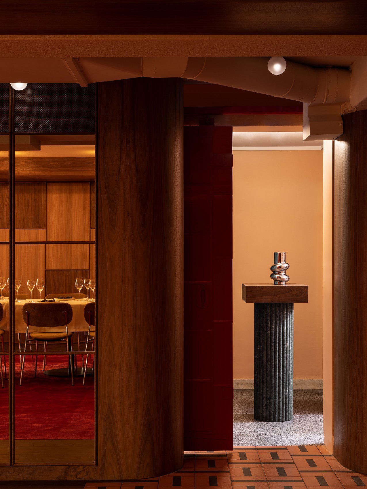 Interior doorway framed by warm wood panels and terracotta tiles, showing a fluted dark stone pedestal with a polished