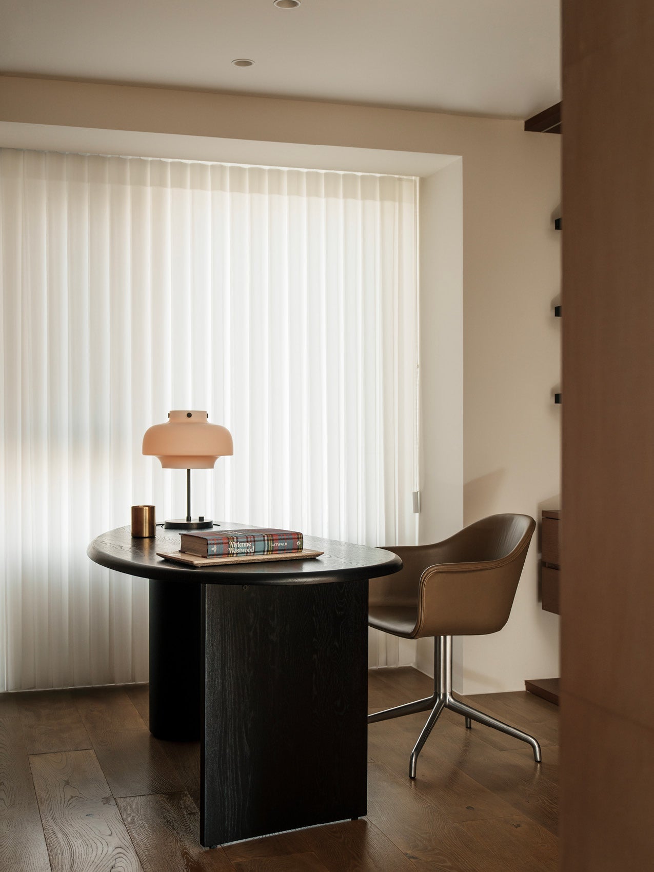 Black oval wooden table with a matte peach glass lamp, stacked books and brass cup beside a tan swivel chair, sheer white
