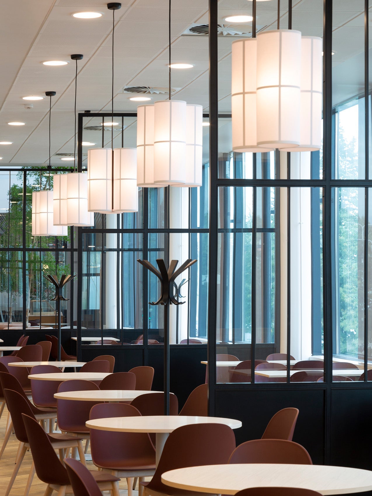 Dining area with round light wood tables and burgundy shell chairs beneath cylindrical white fabric pendant lamps.