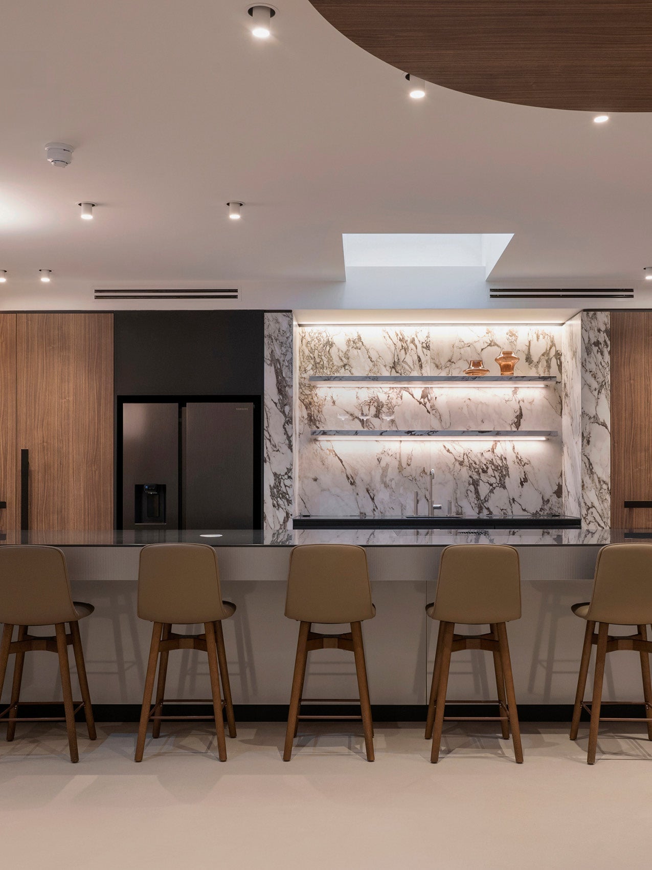 Modern kitchen bar with five beige leather stools, dark marble countertop and veined marble backsplash with backlit shelves