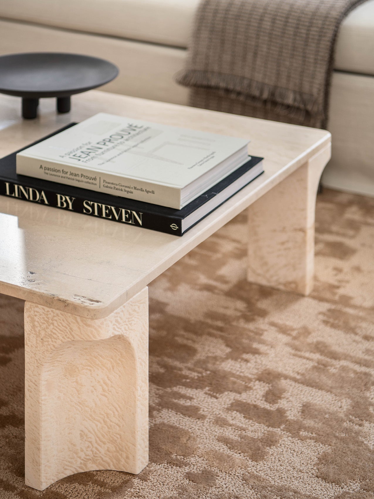 Beige travertine low coffee table with rounded legs, stacked black and white hardcover books and a matte black shallow bowl