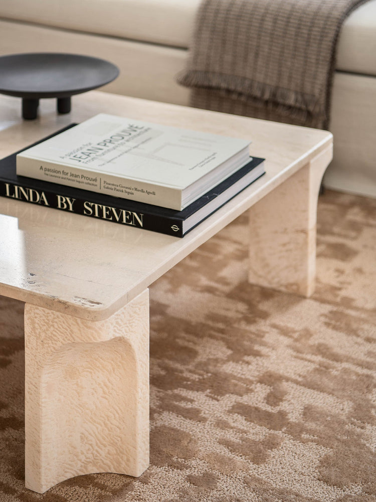 Beige travertine low coffee table with rounded legs, stacked black and white hardcover books and a matte black shallow bowl