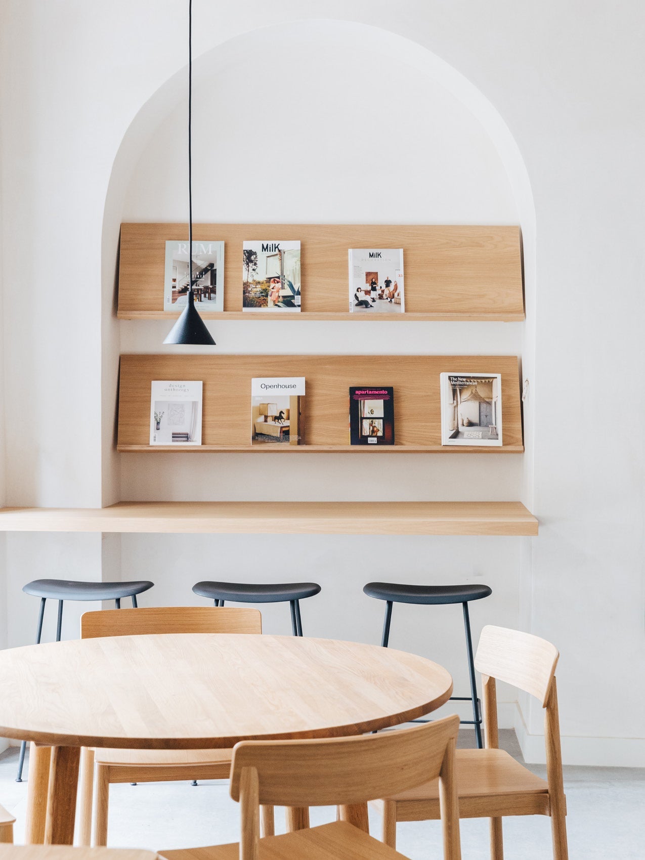 Minimal dining area with round light oak table, matching chairs, three black bar stools, pendant lamp and wood magazine