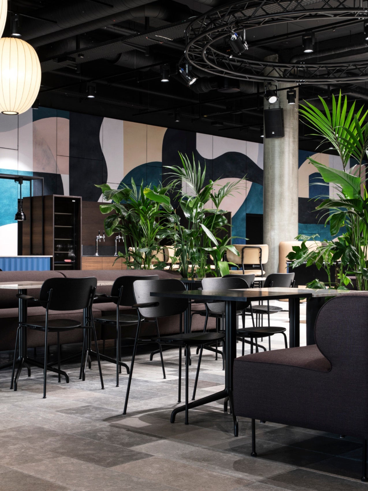 Modern café seating with black metal chairs, dark upholstered benches, round tables and tall green potted plants.