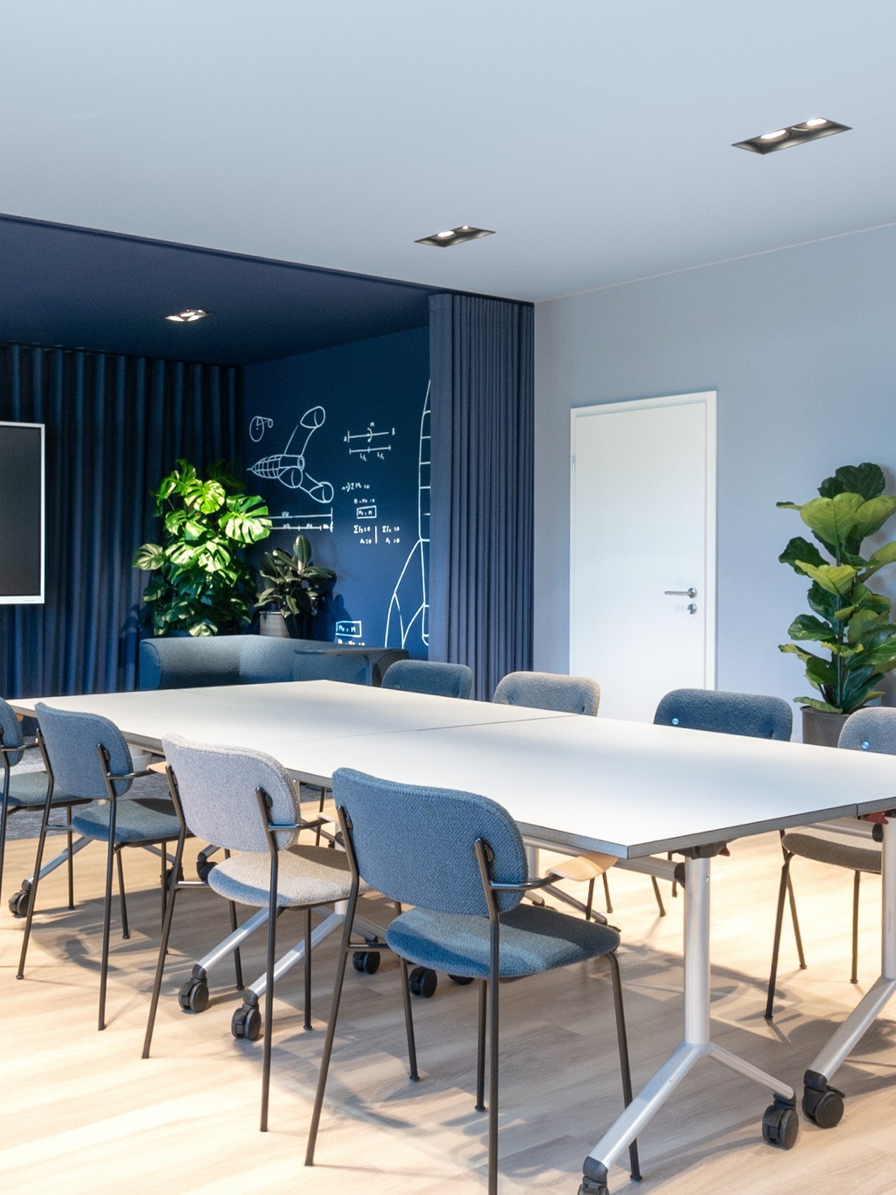 Modern meeting room with white rectangular tables on wheels, gray-blue upholstered chairs, blue accent wall and potted plants