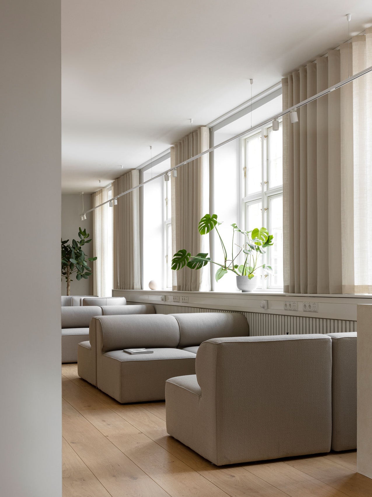Minimal Scandinavian lounge with beige modular sofas, light oak floor, tall cream curtains and a potted monstera on the