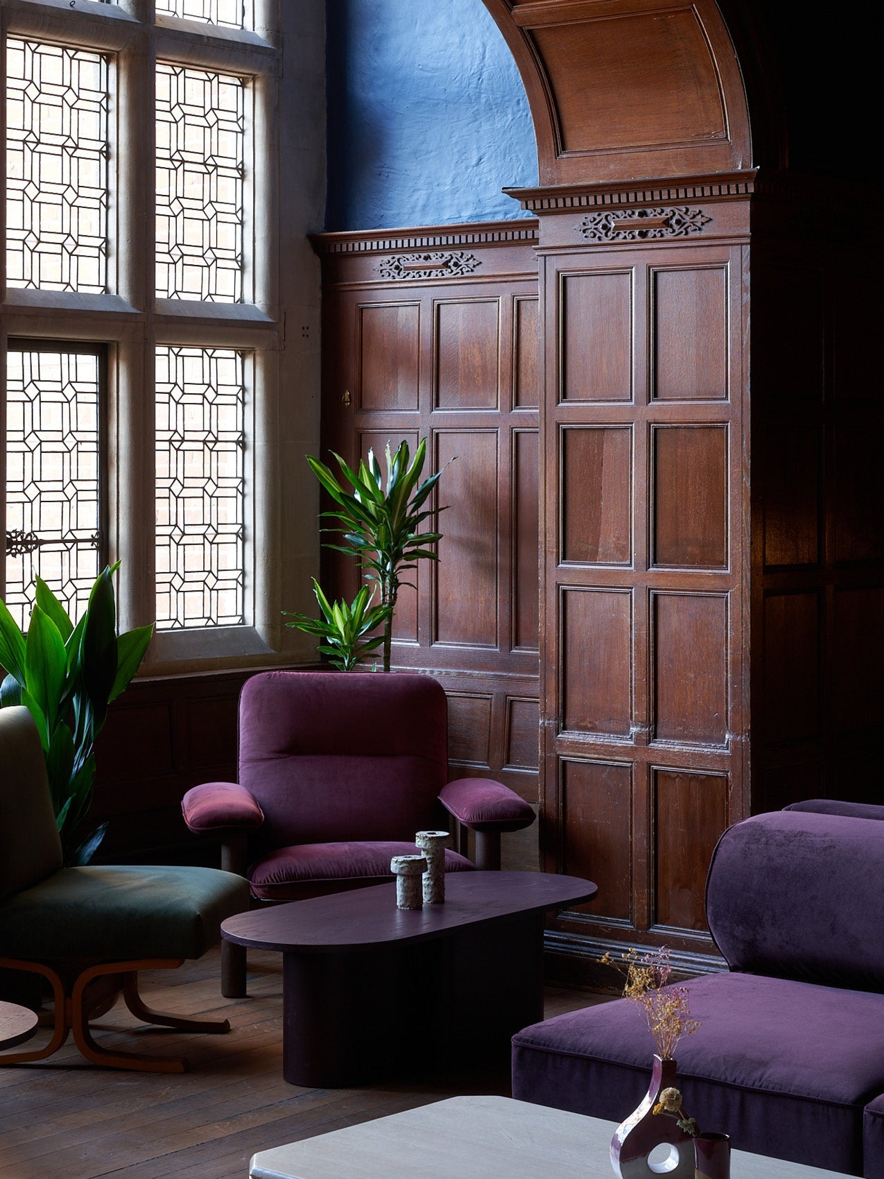 Velvet plum lounge chairs and ottoman with dark wood coffee table against carved oak paneling and leaded glass windows