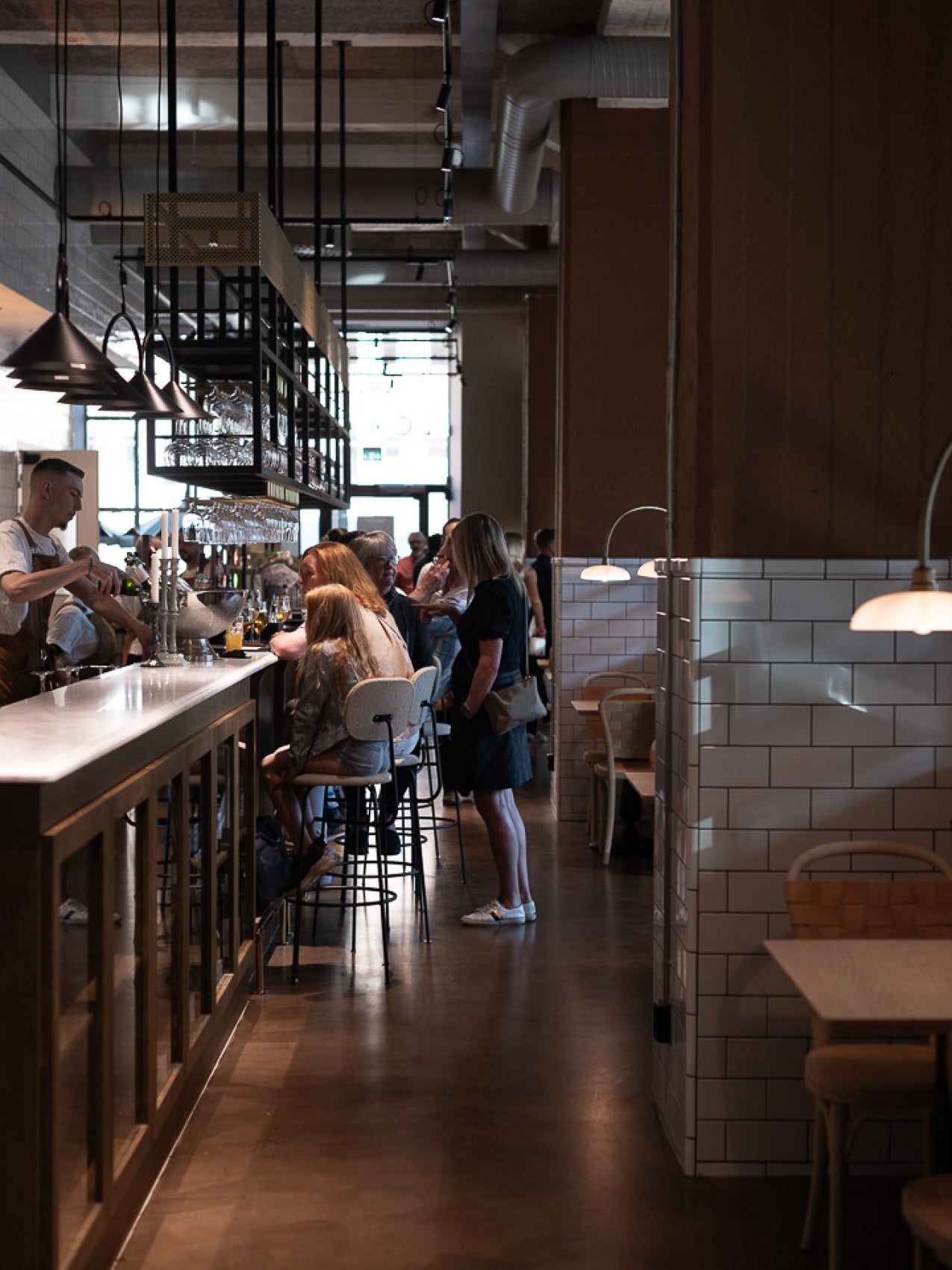 Long marble-topped bar with black metal shelving, pendant lights and patrons seated on light wood and upholstered bar stools