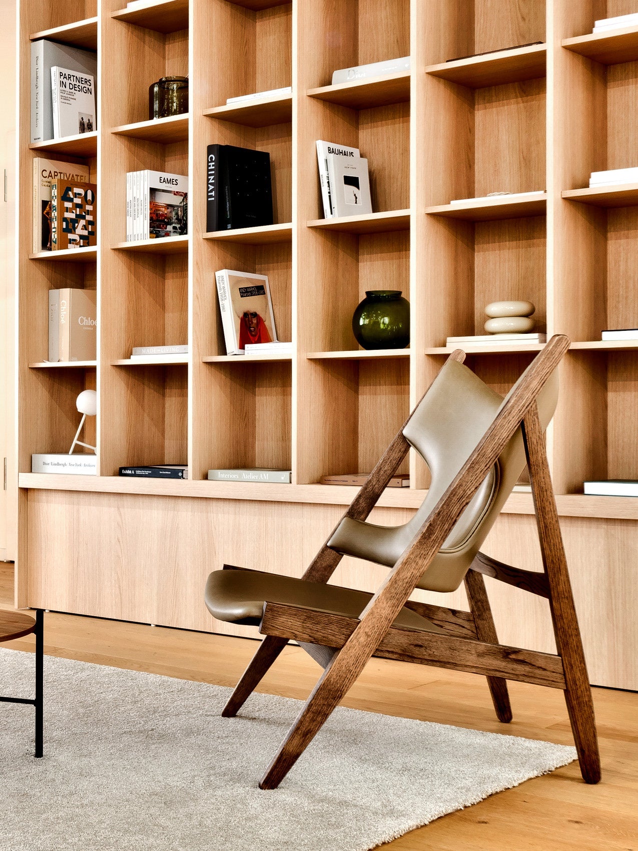 Oak shelving unit with cubed compartments holding books and decor; angled walnut-framed leather lounge chair in foreground.