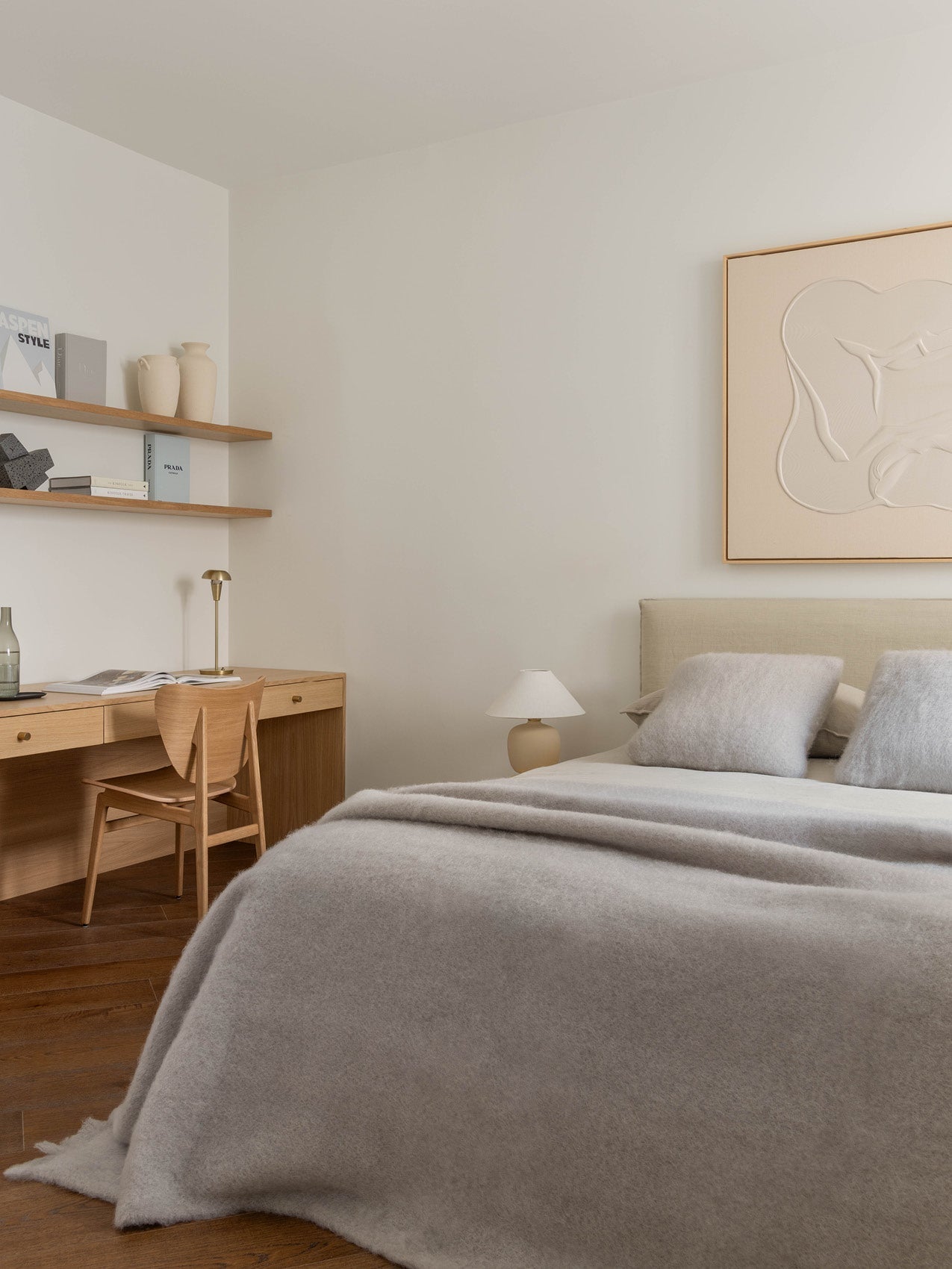 Minimal bedroom corner with beige upholstered bed, soft grey wool blanket and pillows, oak desk and shelving with ceramics.