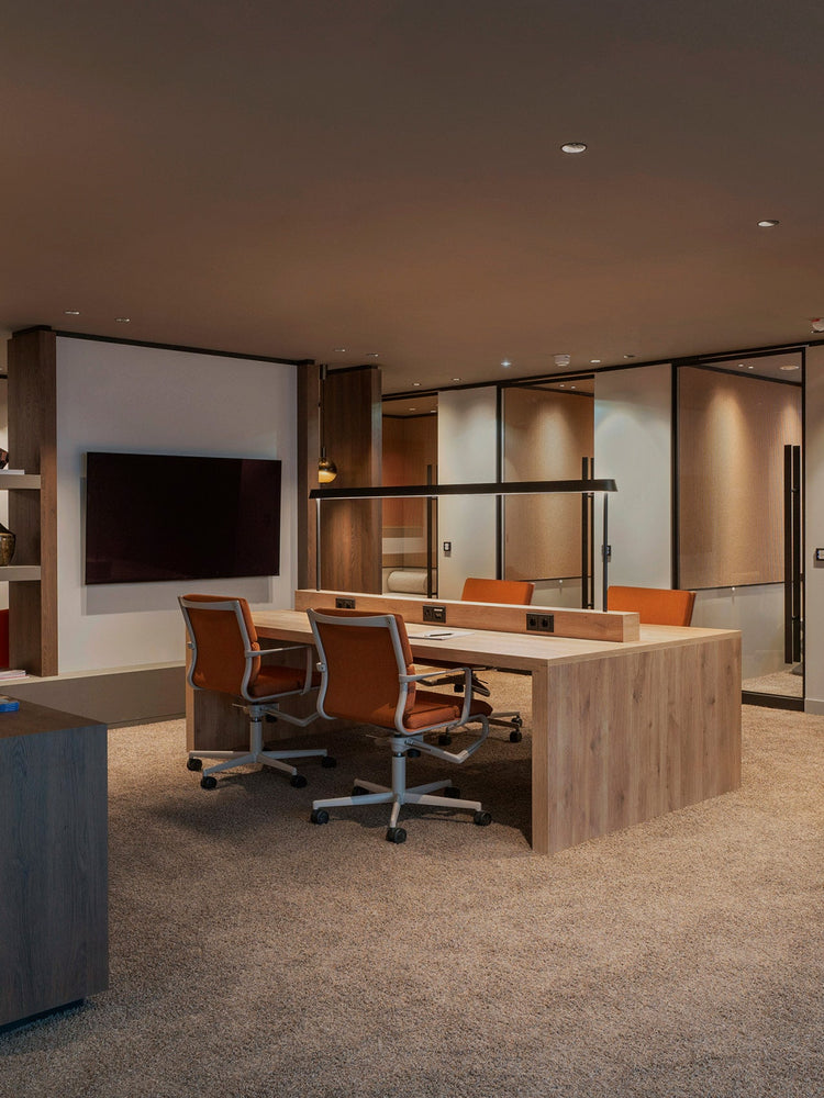 Office workspace with a long light wood desk, integrated power strip, two orange upholstered swivel chairs and horizontal