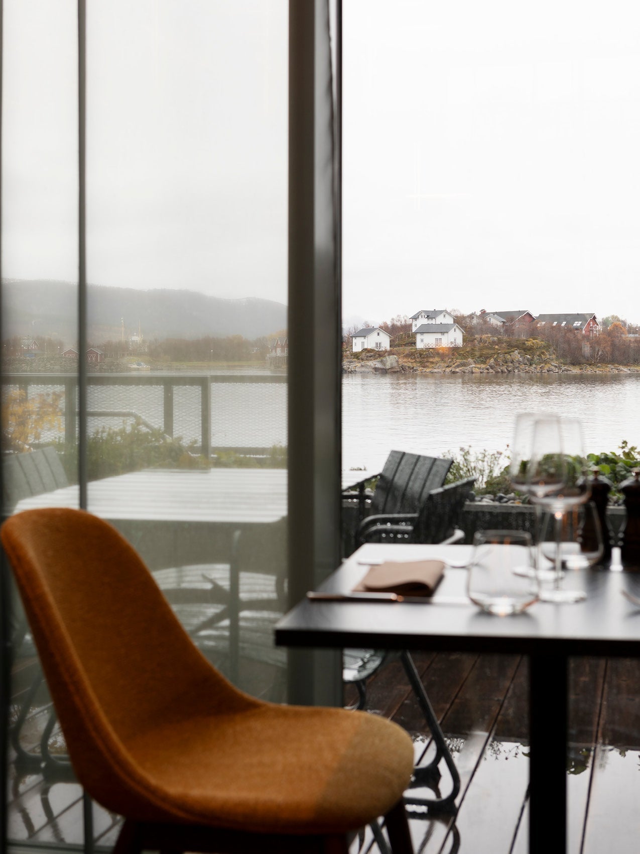 Interior view of a rust-coloured upholstered chair and dining table by a glass door, looking out to a calm waterfront and