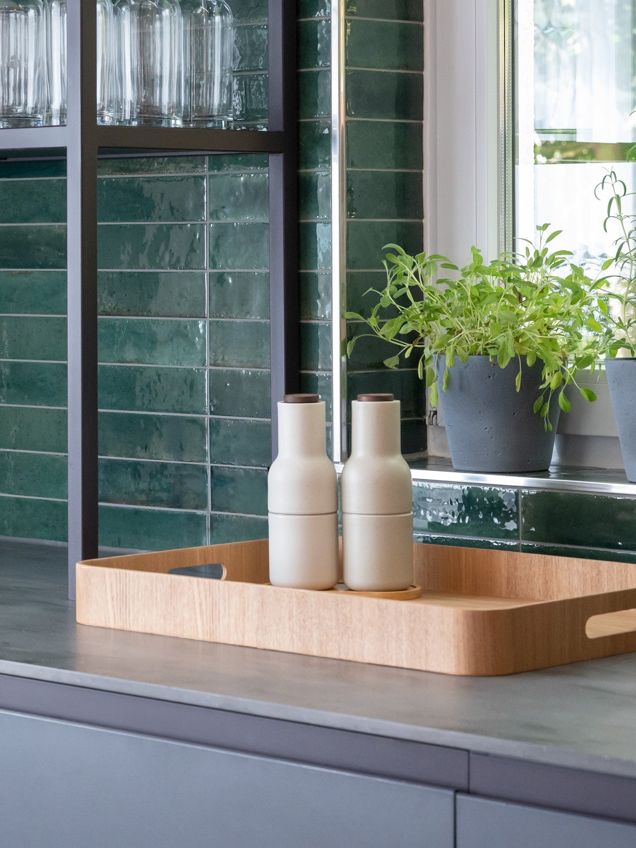 Oak serving tray with two matte beige ceramic bottles on a grey countertop, green subway tile backsplash and potted herbs.