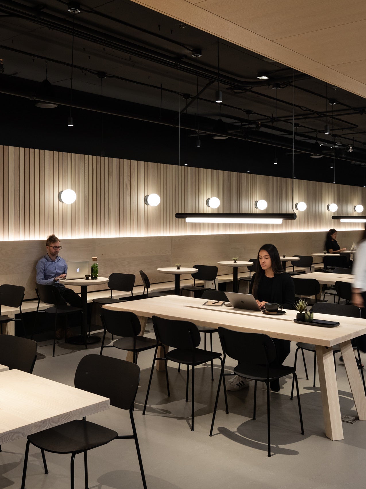 Minimal café interior with light oak communal tables, black chairs, round wall lights and pendant linear lamps, people