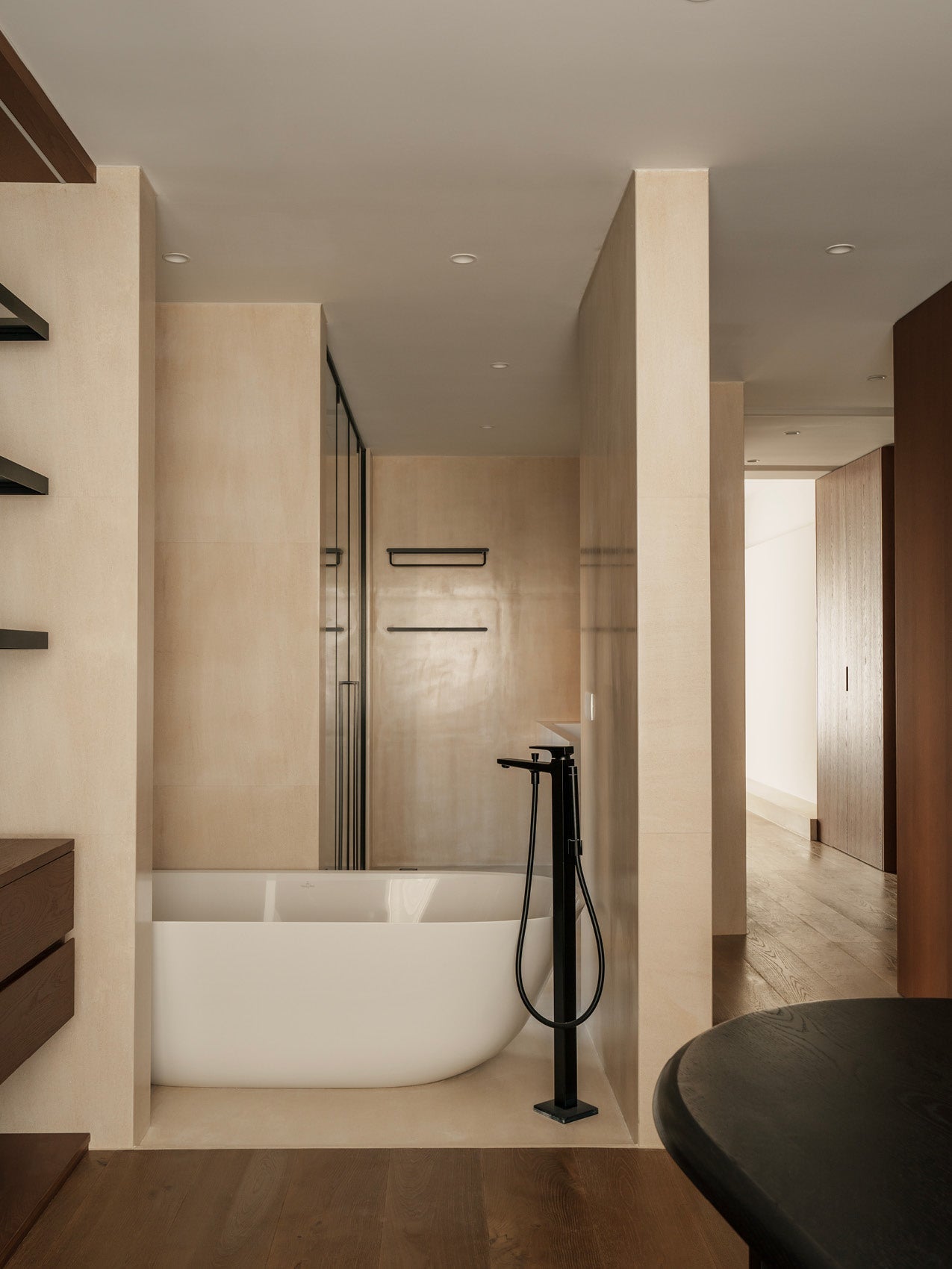 Minimal bathroom with freestanding white oval bathtub, matte black floor-mounted faucet and beige stone walls.