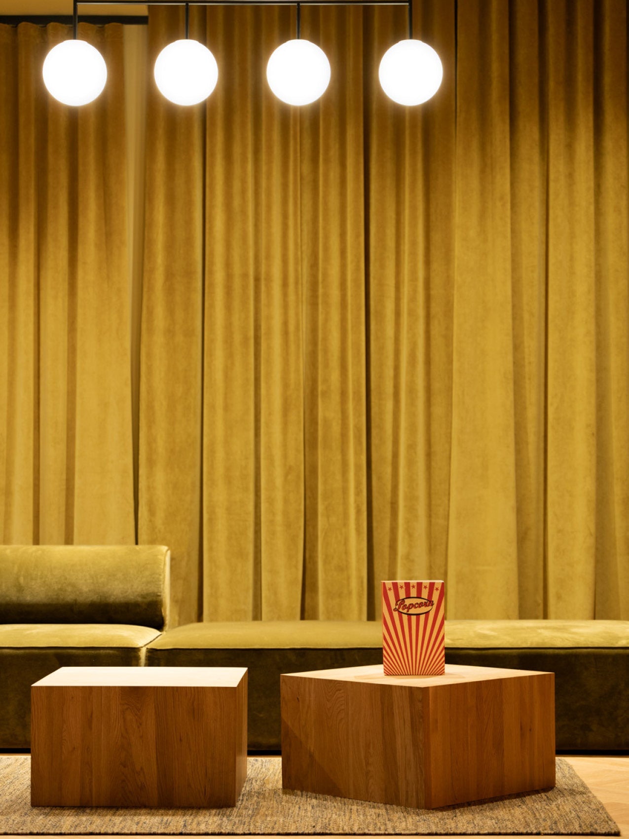 Minimal lounge with mustard velvet curtain, olive green sofa, three round pendant lights and wooden block tables, popcorn