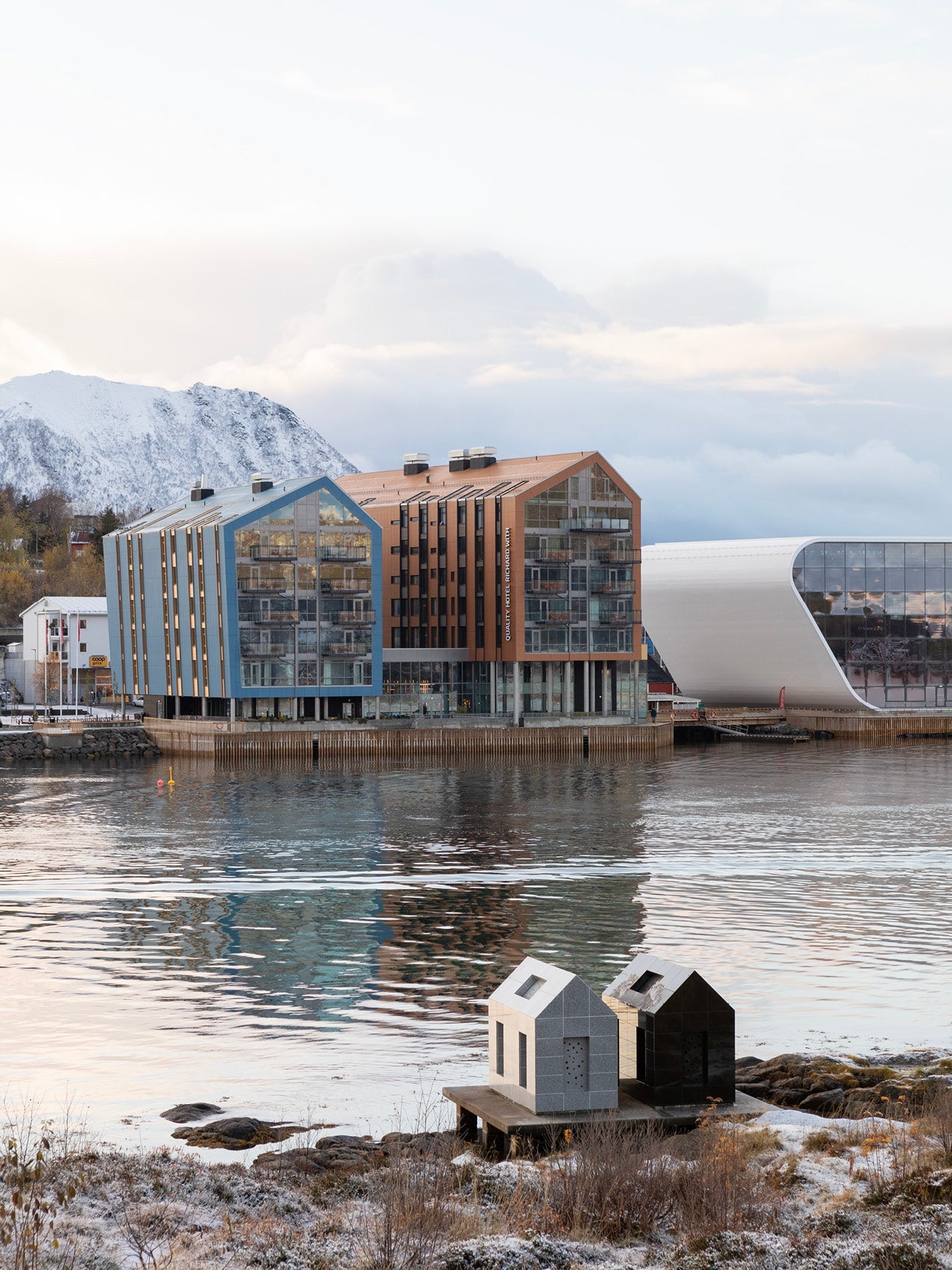 Waterfront modern buildings with glass facades and copper-blue gabled roofs reflected in calm water, snowy shoreline and
