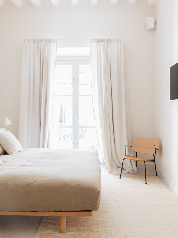 Minimal beige bedroom with linen bed, floor-to-ceiling cream curtains and a light wood chair with black metal legs.