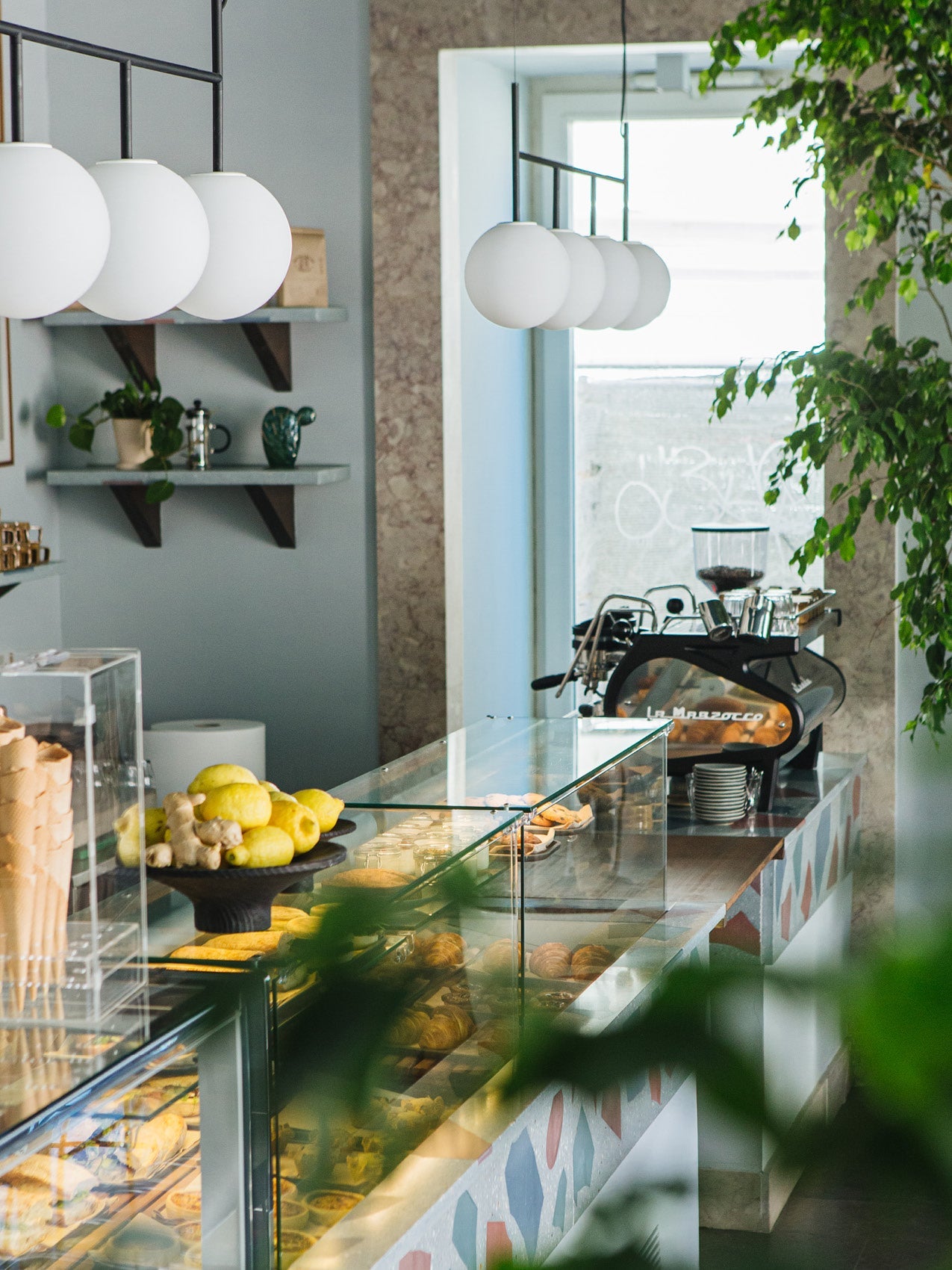 Cafe counter with glass display of pastries, lemons on a black pedestal, white globe pendant lights and espresso machine