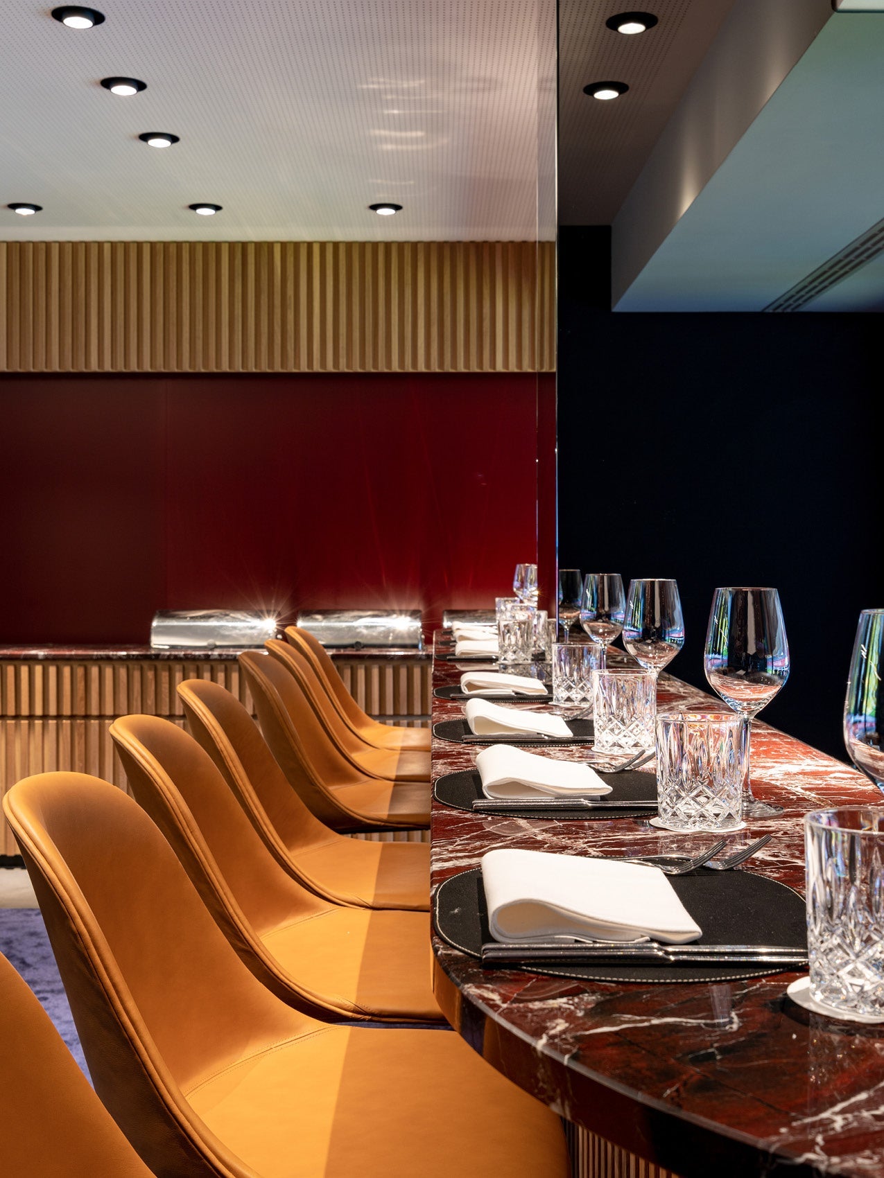 Long marble bar with crystal glassware and white napkins, lined with tan leather curved stools and wooden slatted wall.