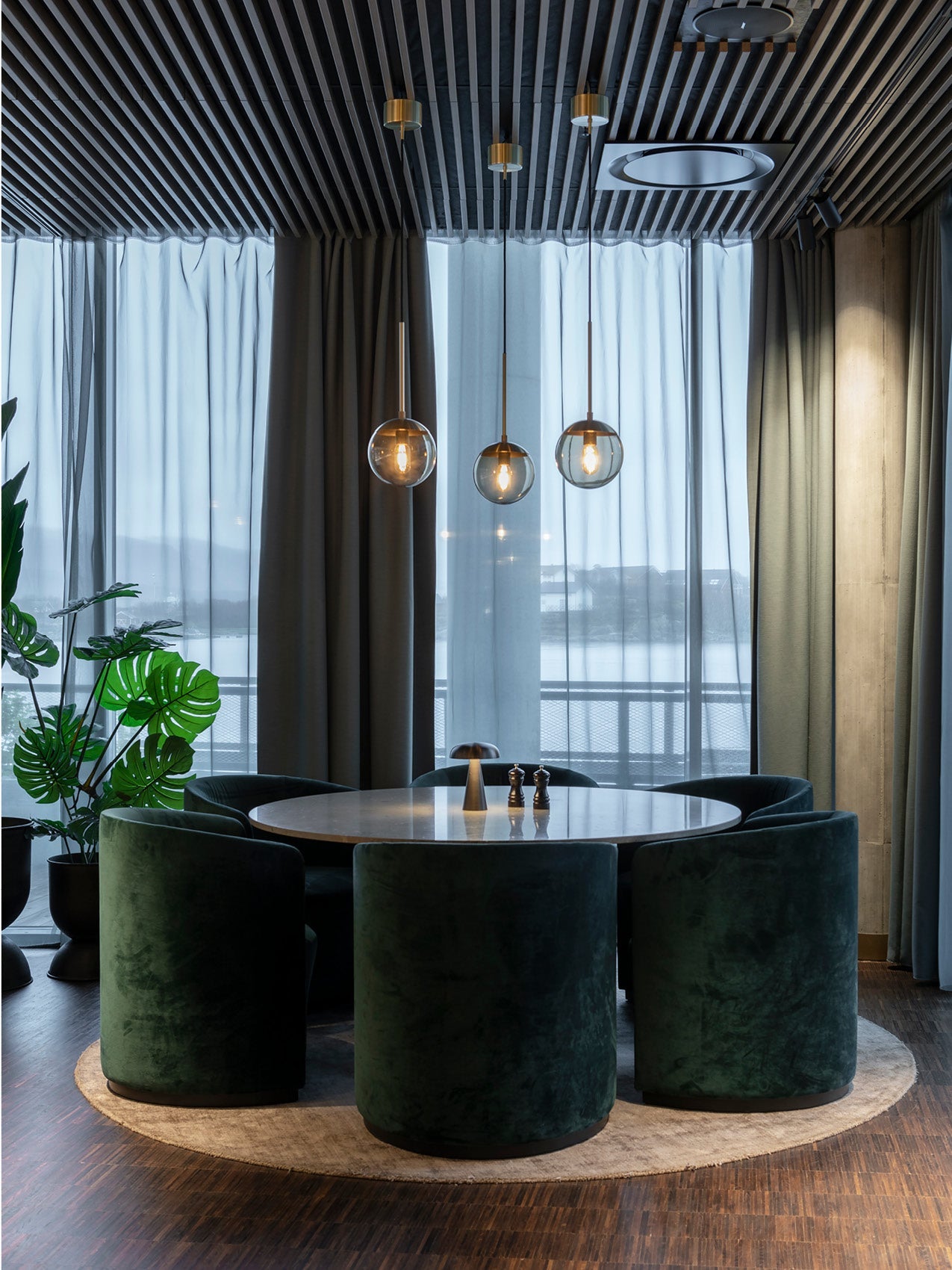 Round marble dining table surrounded by deep green velvet barrel chairs, glass globe pendant lights and sheer curtains.