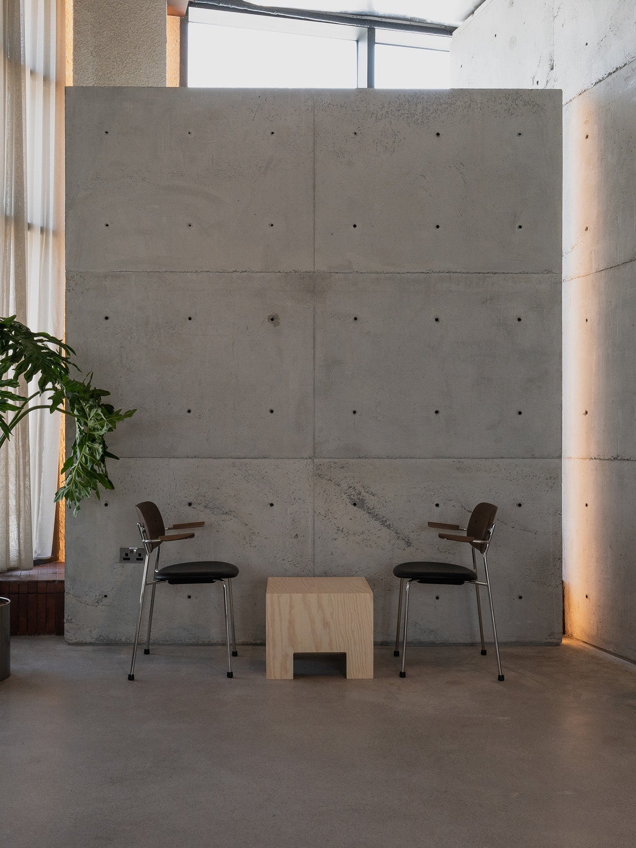Minimal interior with two black leather and wood chairs facing a light plywood cube table against a raw concrete wall