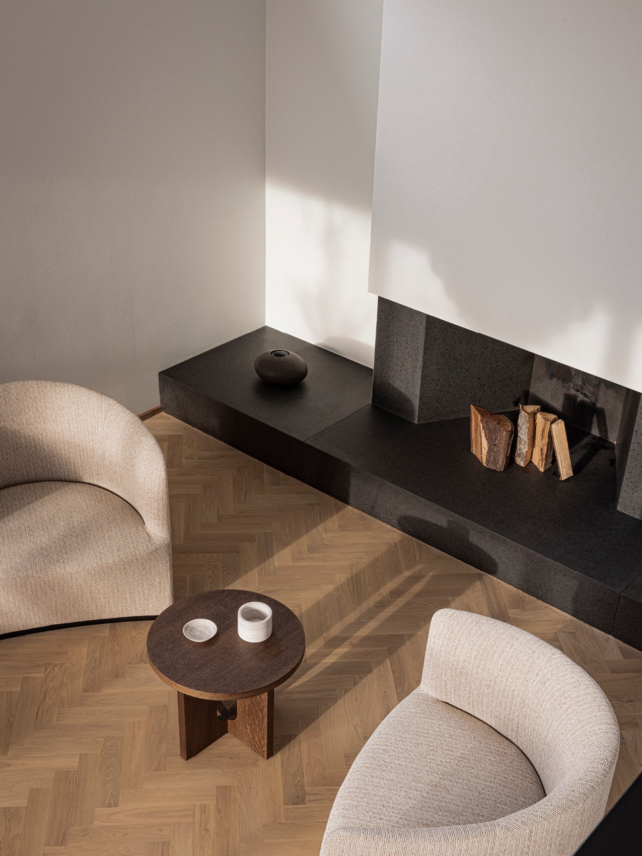 Minimal living room corner with curved beige upholstered armchairs, round dark wood side table, black stone hearth and