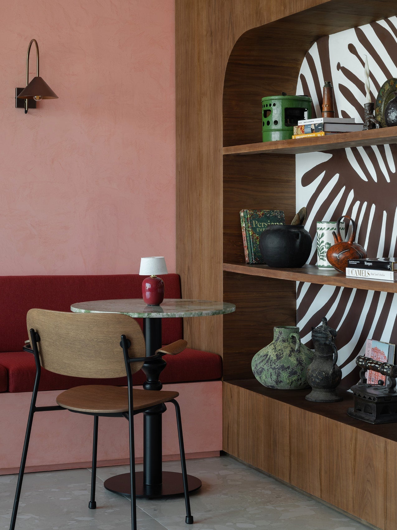 Small round marble-top table with a red ceramic lamp, wooden chair, pink plaster wall and built-in wood shelves with