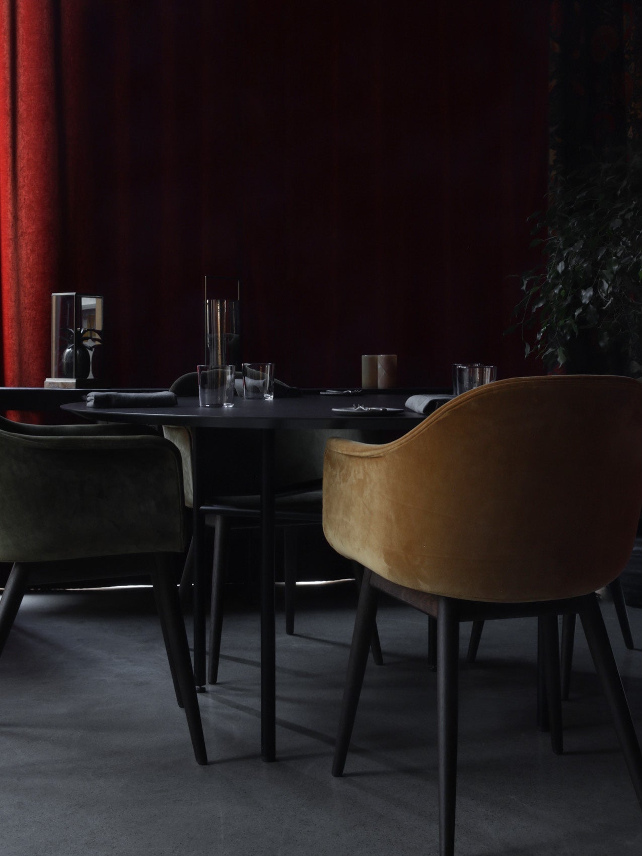 Round black dining table with glassware and velvety curved chairs in mustard and dark green against deep red curtains
