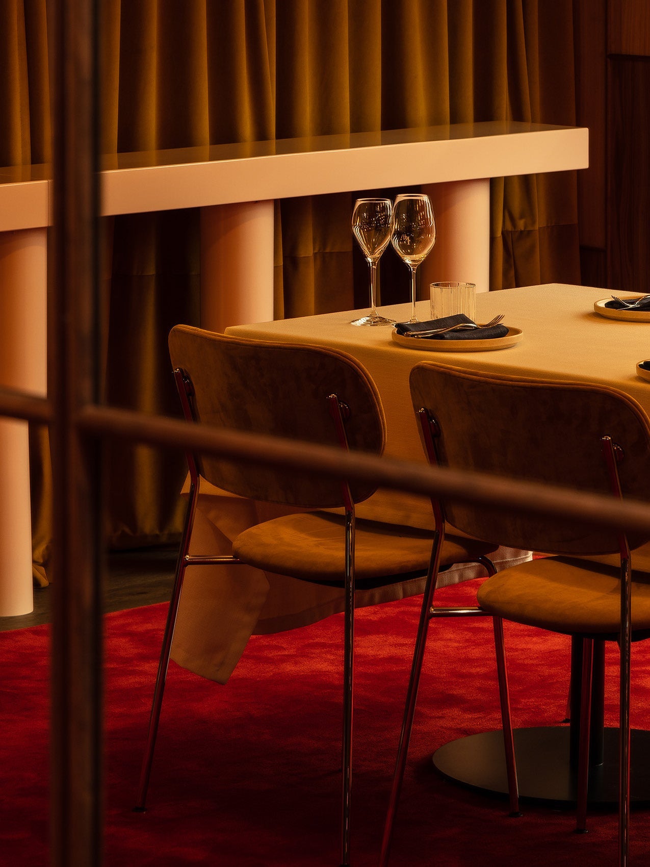 Dining table set with beige tablecloth, two velvet brown chairs, wine glasses and plates on red carpet background