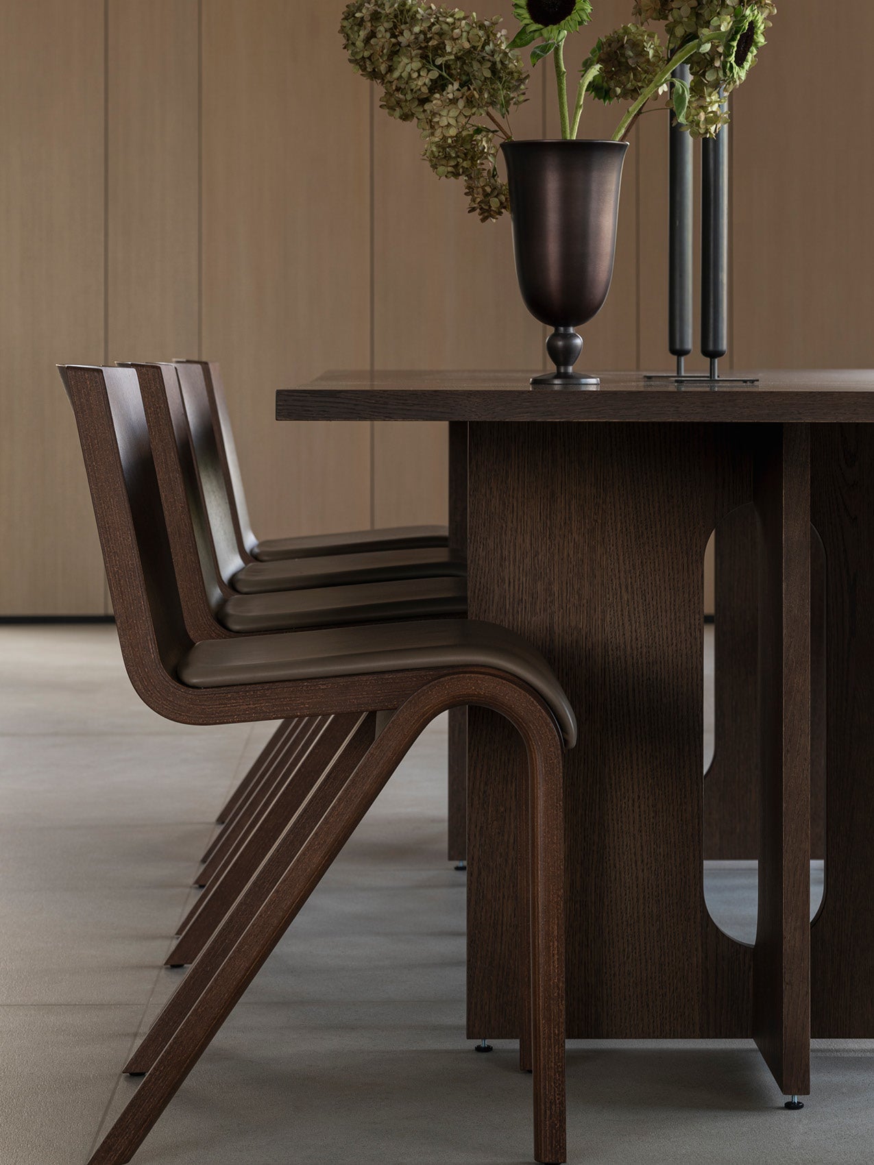 Row of curved dark walnut dining chairs tucked under a matching dark wood table with bronze vase and dried flowers on top