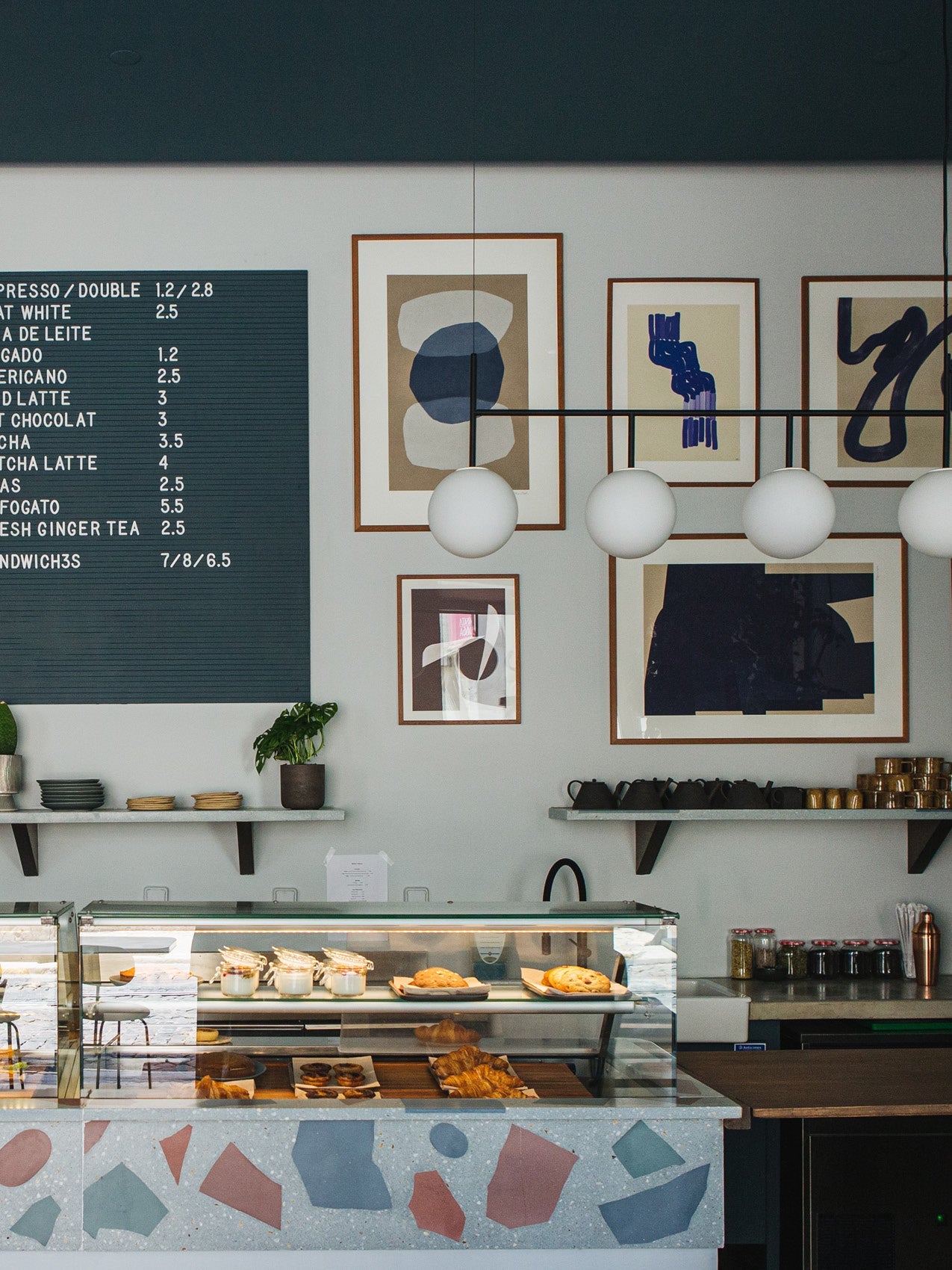 Minimal café counter with terrazzo front, glass pastry case, hanging white globe lights and framed abstract art on pale wall