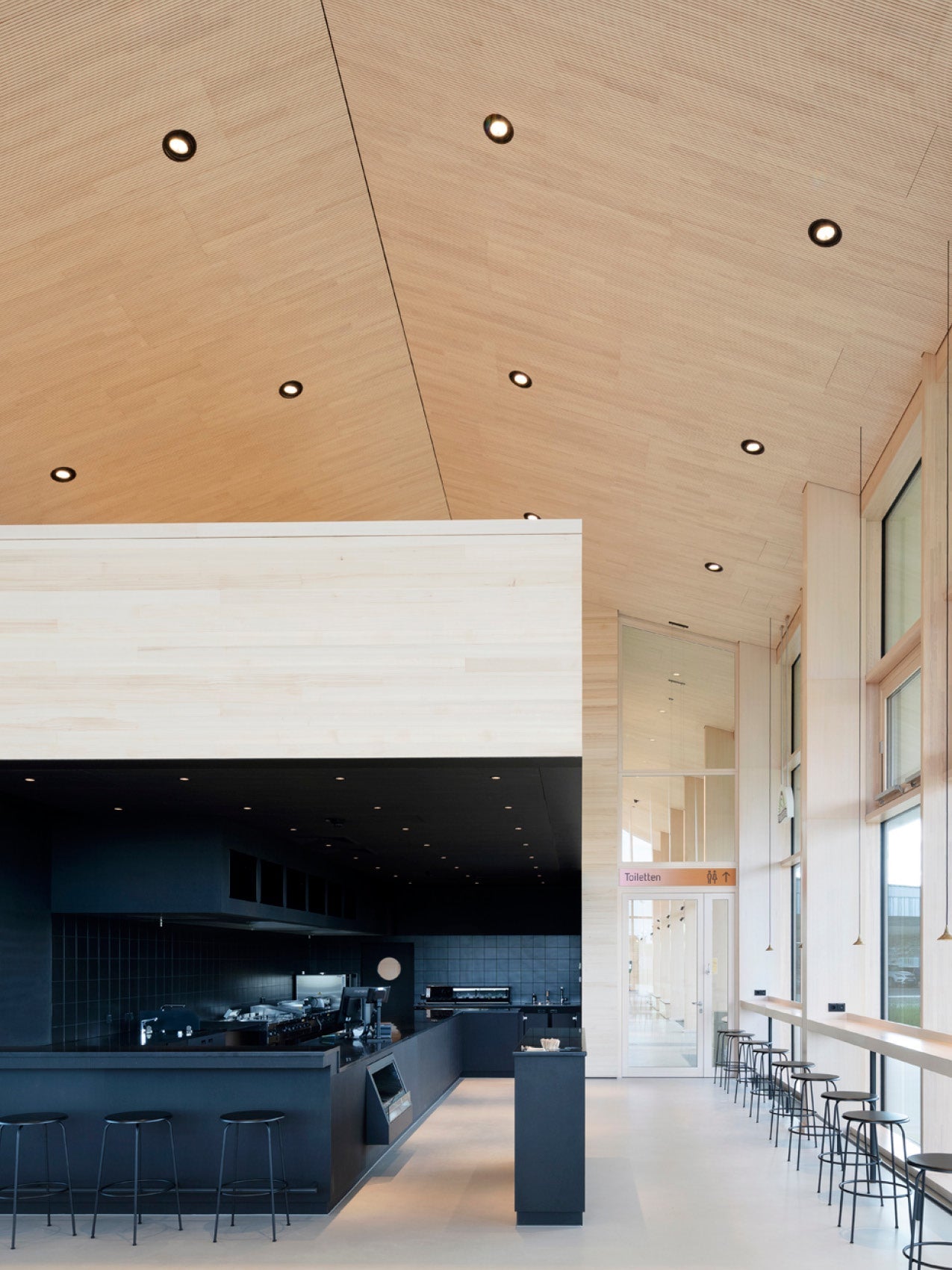 Minimalist café interior with light wood vaulted ceiling, black service counter and row of black metal stools by tall windows