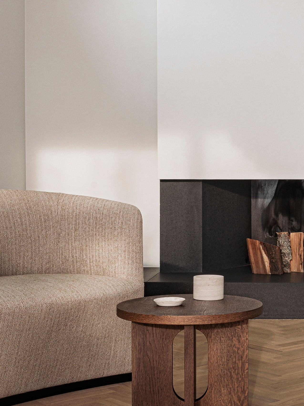Beige curved fabric sofa beside dark stone fireplace with stacked firewood and round wooden side table with decor