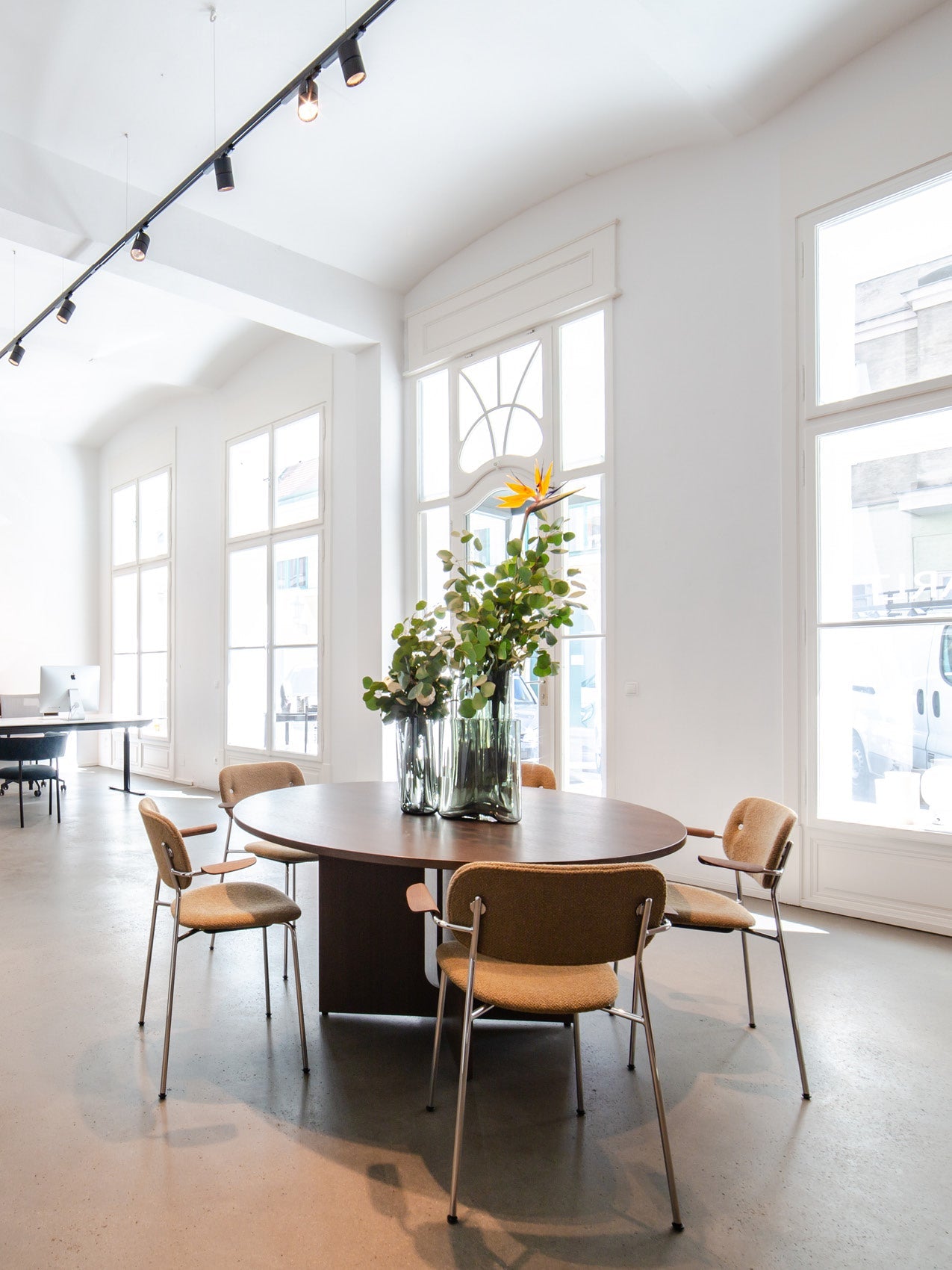 Bright white room with a dark round wooden table, tan upholstered chairs and tall glass vases of green foliage and orange