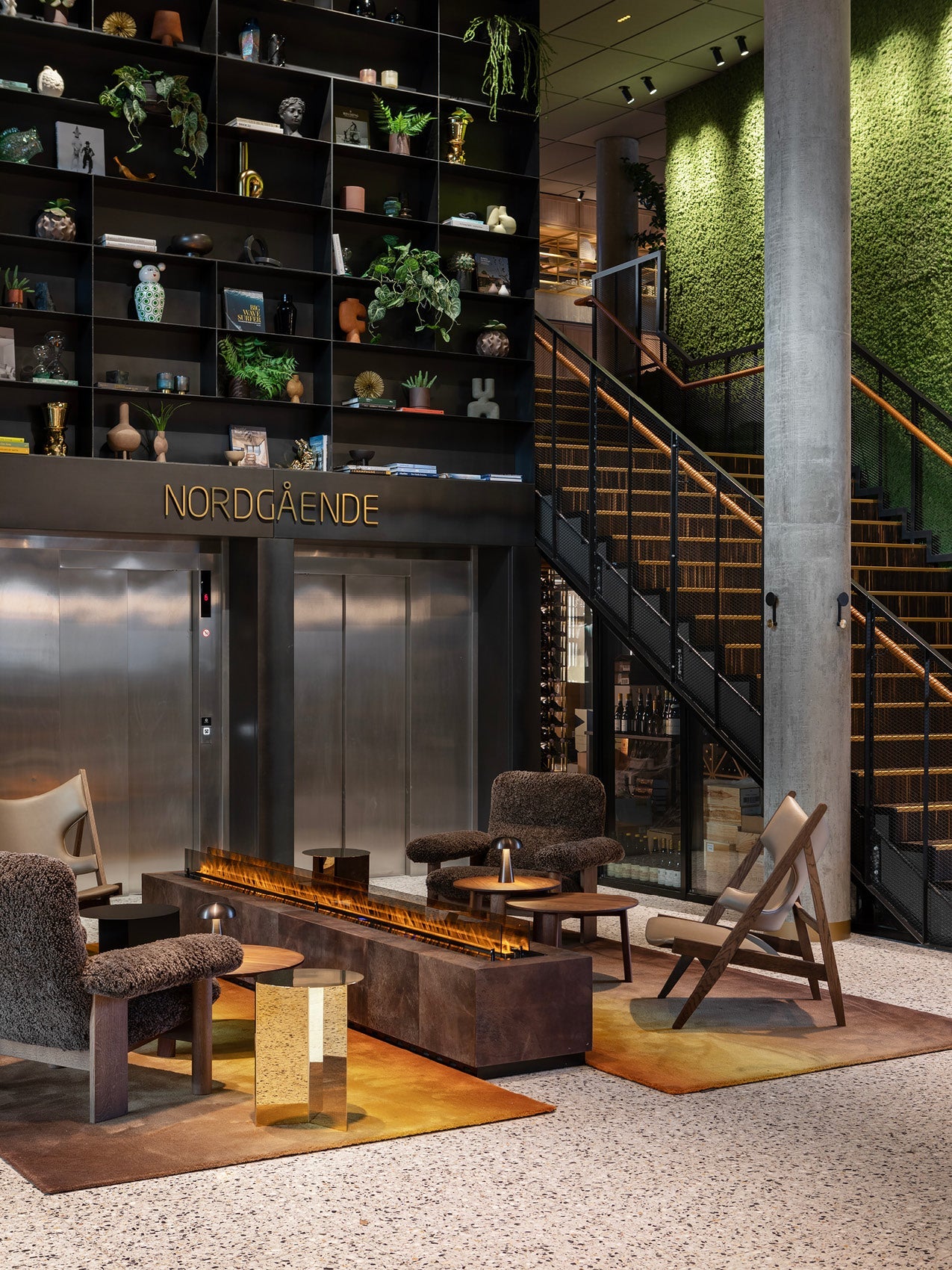 Contemporary hotel lounge with brown boucle armchairs, low rust-colored fireplace, brass side table, shelving wall and green