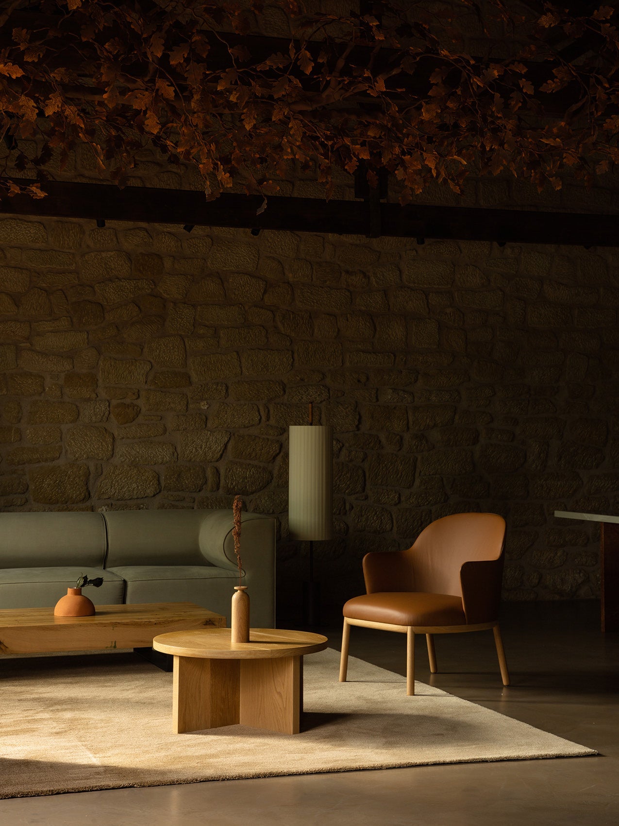 Minimal living room with stone wall, green sofa, tan leather lounge chair, round oak coffee tables and muted pendant lamp.