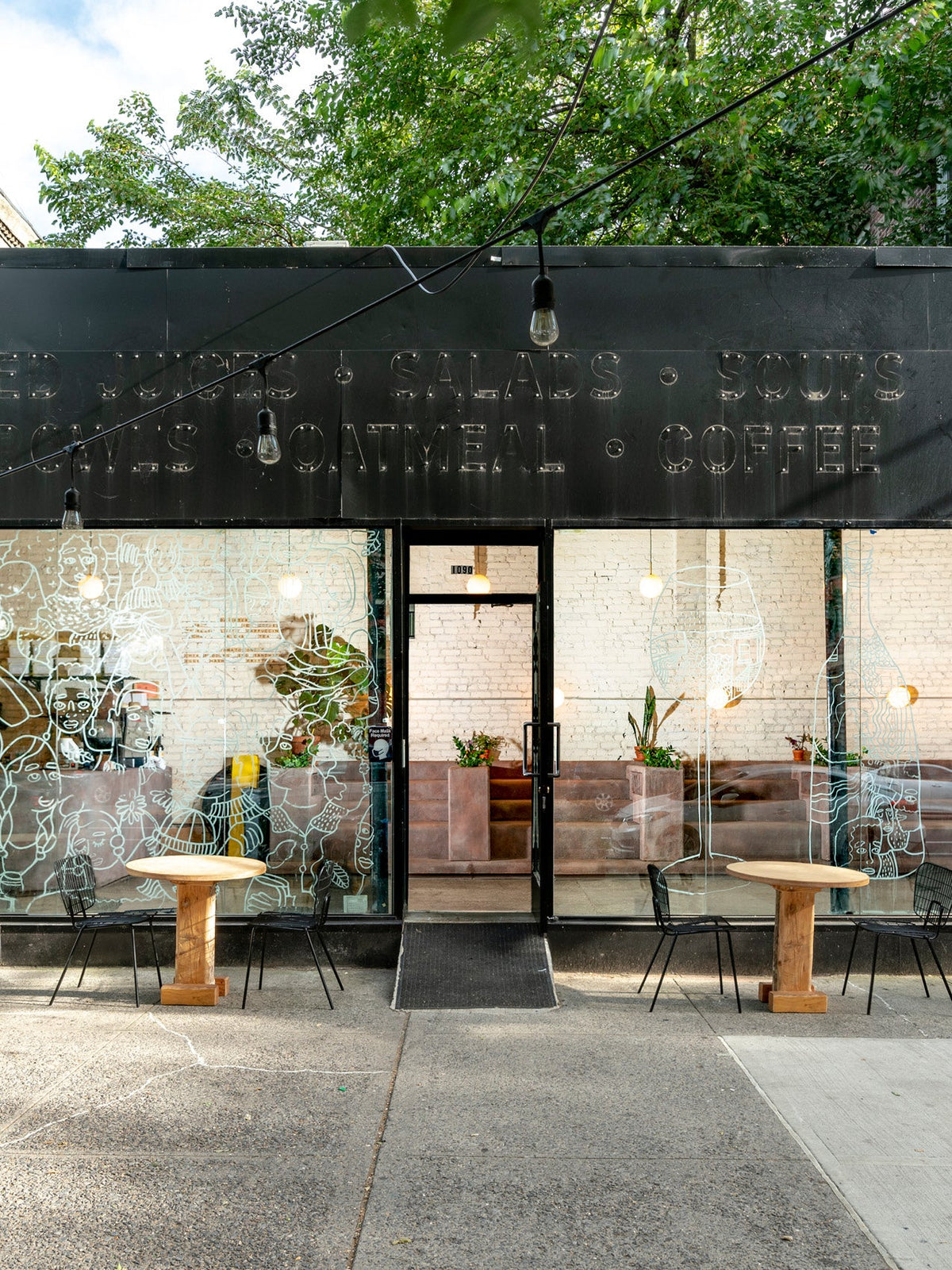 Black wire chairs and wooden cafe tables set outside a storefront with white window illustrations and a dark sign.