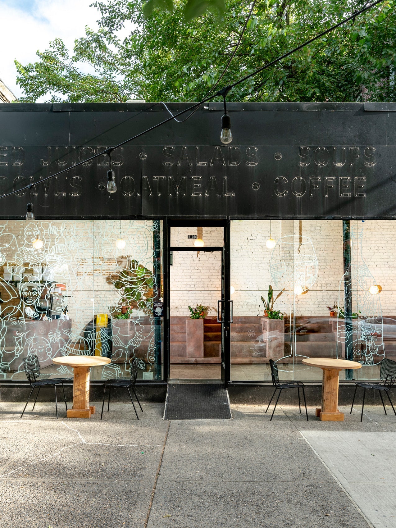 Black storefront with glass windows and white mural drawings, two round wooden café tables and wire chairs outside.