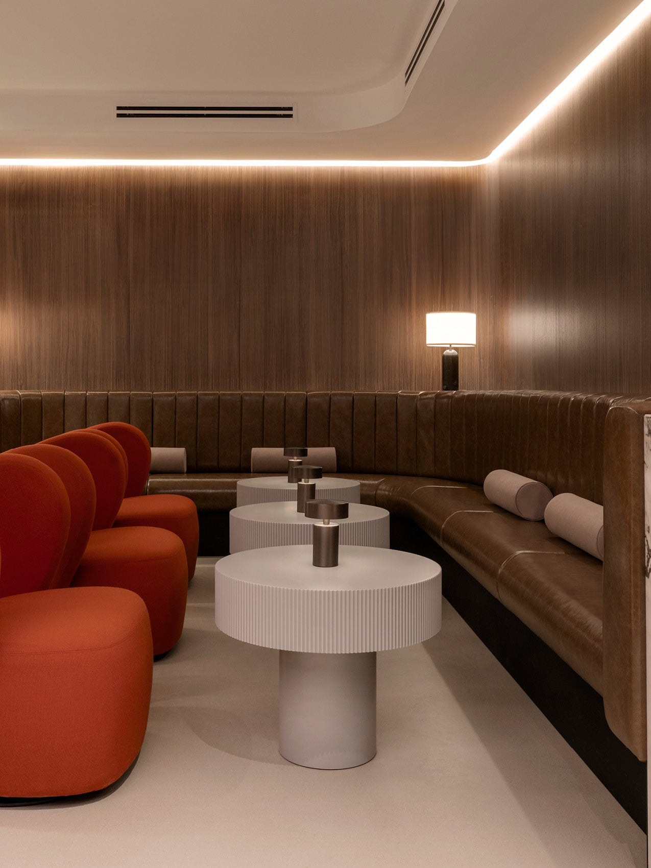 Minimal lounge with curved brown leather banquette, orange upholstered chairs, round ribbed white side tables and warm lamp