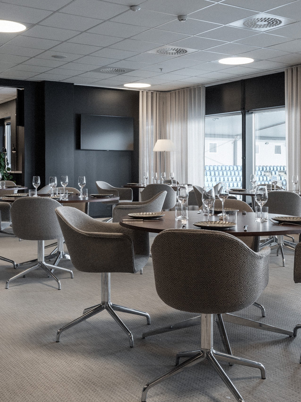 Dining room with round dark wood tables and grey upholstered swivel chairs, set with glassware and plates, soft neutral